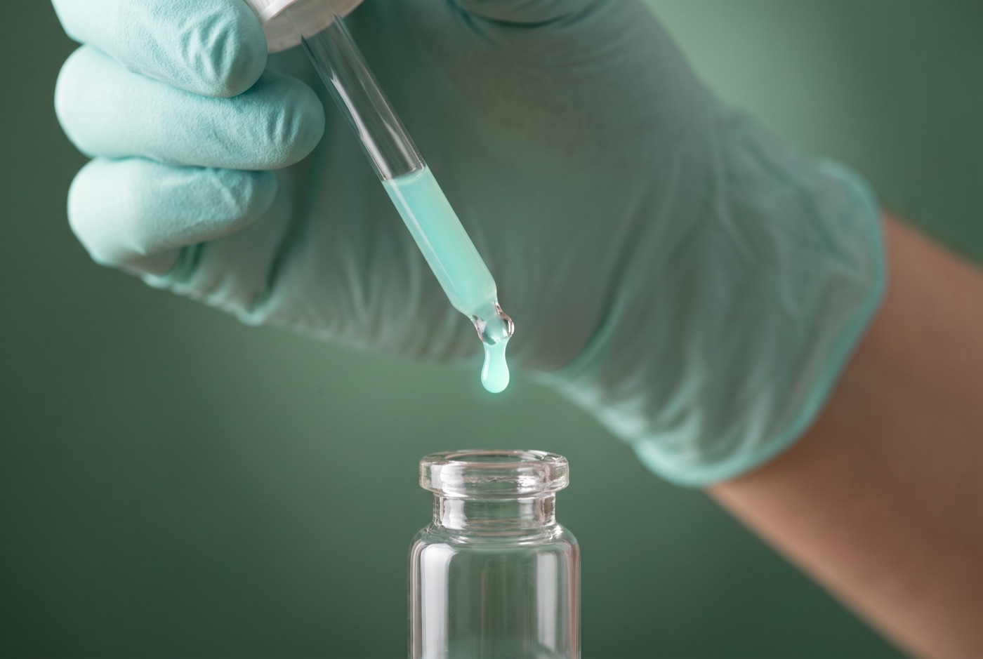A gloved hand dispensing a single teal drop from a pipette into a vial