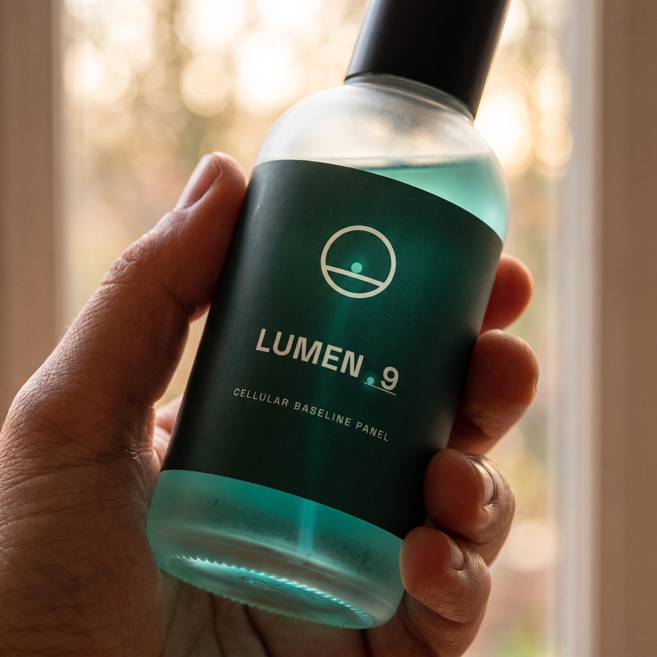 A hand holding the labeled LUMEN-9 concept bottle against warm window light, label sharp