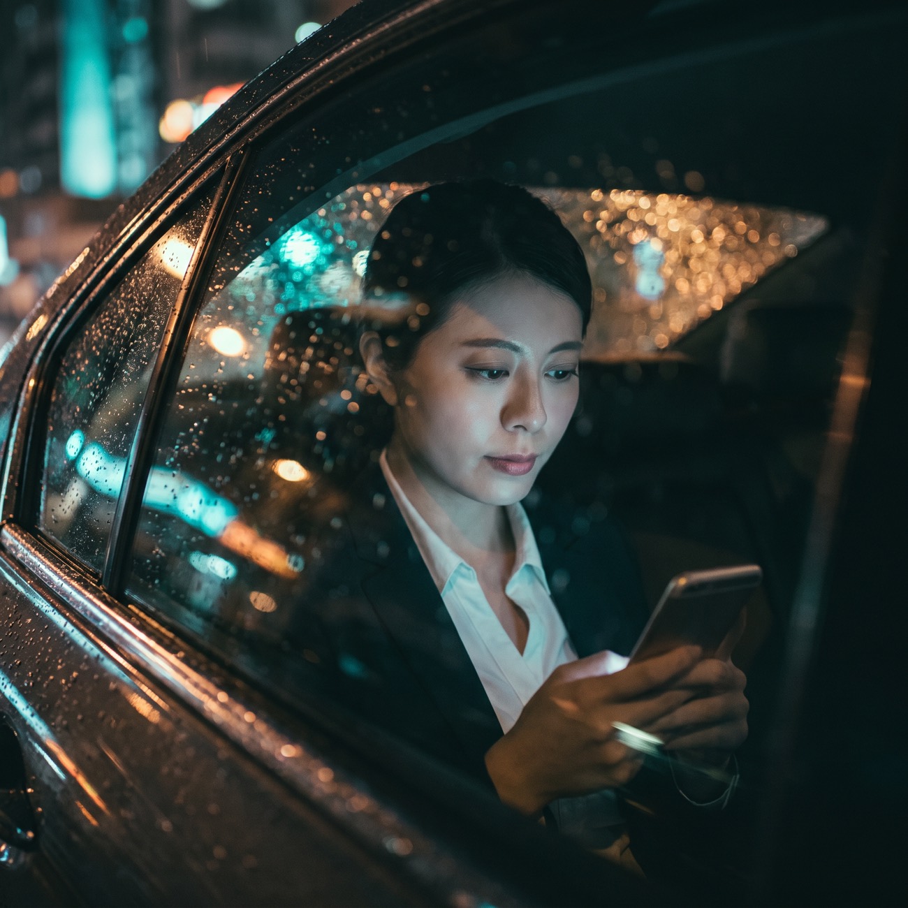 An investor reading her phone in the back of a car at night, city lights through the rain-flecked window