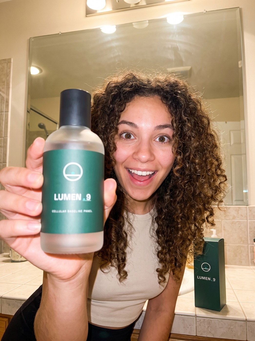 Creator-style phone photo: a young woman excitedly holds the labeled LUMEN-9 concept bottle to the camera in her bathroom